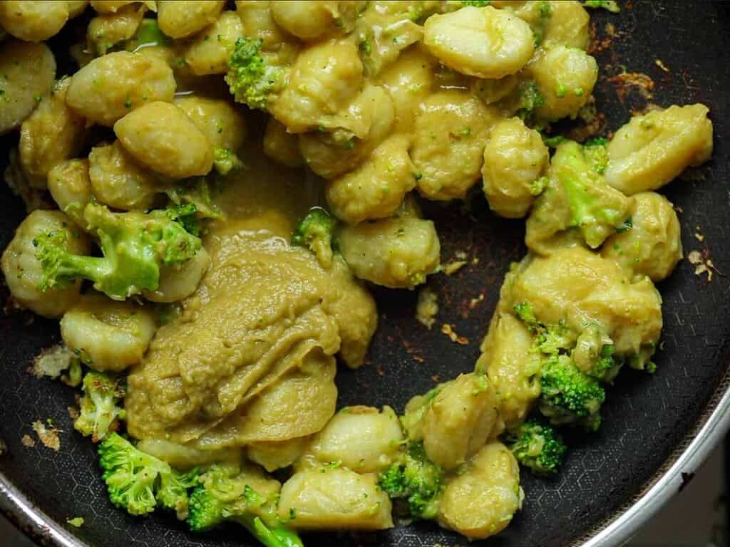 A dish of gnocchi with broccoli in a creamy sauce is cooked in a black skillet. The sauce appears rich and covers the gnocchi and broccoli pieces, creating a savory, visually appealing meal.